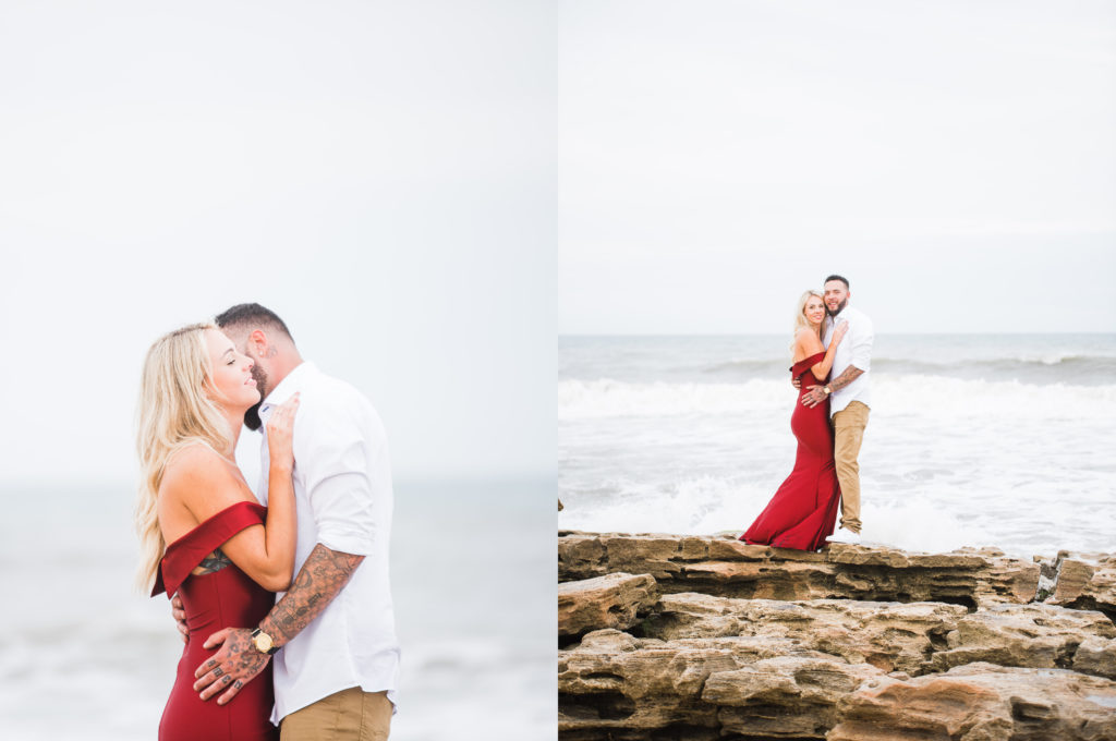 Beach engagement session in palm coast. couple stand on rocks, bride is wearing a red form-fitting dress. photo by Chabeli Woolsey of Black Tie & Co www.btcweddings.com